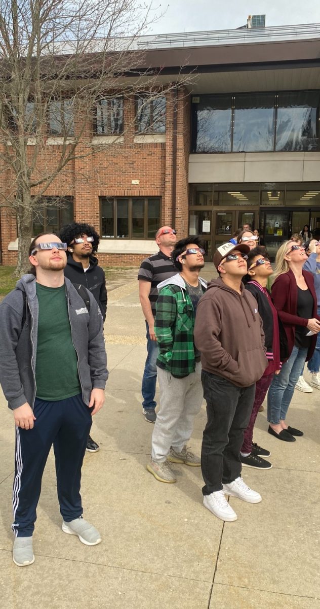 students and staff wearing eclipse glasses look up at the eclipse in front of the Peconic Building